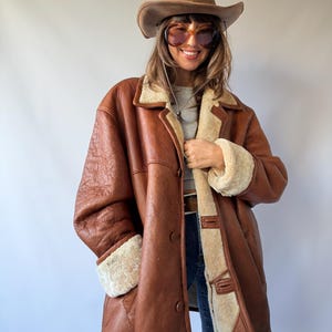 Könnte beinhalten: Ein brauner Ledermantel mit dickem, cremefarbenem Lammfellfutter, ausgestattet mit einem breiten Kragen, langen Ärmeln mit Umschlagbündchen und einer Knopfleiste. Der Mantel wird mit einem braunen Cowboyhut und einer getönten Sonnenbrille getragen.