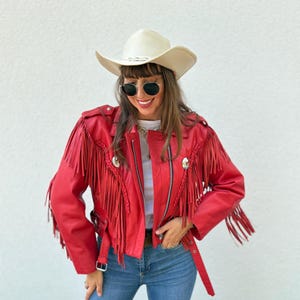 Op de afbeelding: Rode leren jas met franjes, een zilveren rits en decoratieve knopen. De jas wordt gedragen met een blauwe spijkerbroek en een cr&egrave;mekleurige cowboyhoed. De persoon draagt ook een zonnebril.