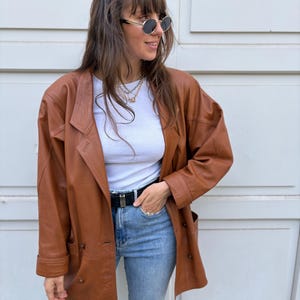 May include: A brown leather blazer with a notched collar, long sleeves, and a double-breasted button closure. The jacket is worn over a white ribbed crew neck top, light blue denim jeans, and a black belt with a silver buckle.