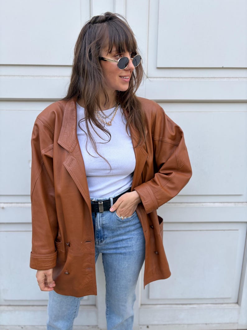 May include: A brown leather blazer with a notched collar, long sleeves, and a double-breasted button closure. The jacket is worn over a white ribbed crew neck top, light blue denim jeans, and a black belt with a silver buckle.