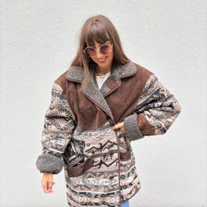 Può includere: Un cappotto marrone in camoscio e fantasia con un design doppiopetto. Il cappotto presenta un mix di camoscio marrone, tessuto fantasia nelle tonalità del beige, grigio e nero e un colletto e polsini in ecopelliccia. Il cappotto ha due tasche anteriori.
