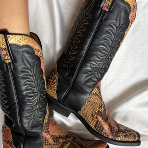 Puede incluir: Un par de botas de vaquero de cuero negro con detalles de piel de serpiente. Las botas tienen costuras decorativas y una puntera puntiaguda. La piel de serpiente es una mezcla de colores marr&oacute;n, naranja y beige.