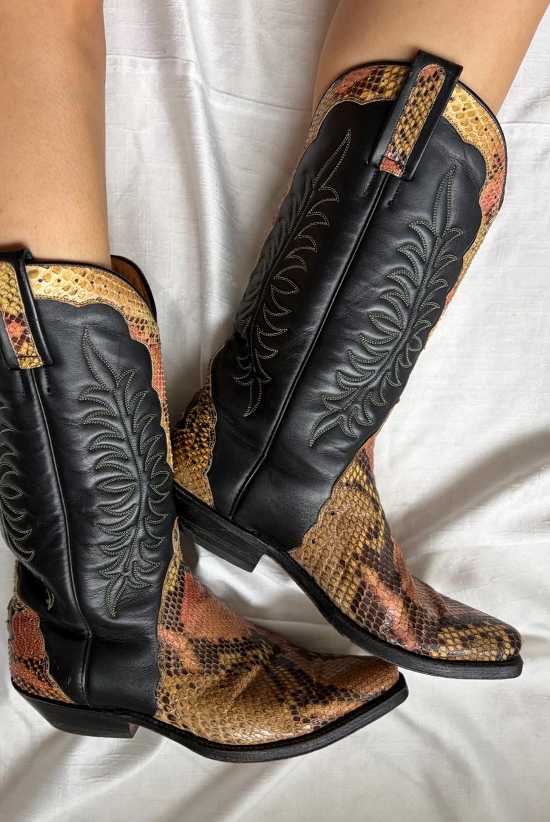 Puede incluir: Un par de botas de vaquero de cuero negro con detalles de piel de serpiente. Las botas tienen costuras decorativas y una puntera puntiaguda. La piel de serpiente es una mezcla de colores marr&oacute;n, naranja y beige.