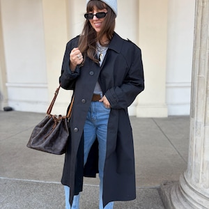 Pode incluir: Um trench coat preto com fecho de botão, usado com jeans azuis e botas de camurça castanhas. Uma touca cinza clara e óculos de sol pretos completam o look. Uma bolsa de balde castanha com estampa de logotipo é carregada no ombro.