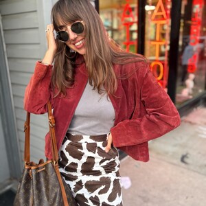 May include: A woman wearing a red suede jacket, gray top, and a cow print mini skirt. She is also wearing sunglasses and carrying a brown bucket bag. The background includes a storefront with the word "TATTOO" in red neon.