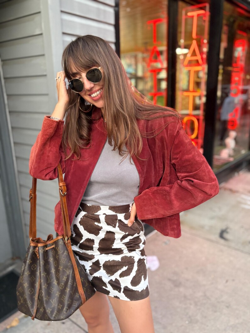 May include: A woman wearing a red suede jacket, gray top, and a cow print mini skirt. She is also wearing sunglasses and carrying a brown bucket bag. The background includes a storefront with the word "TATTOO" in red neon.