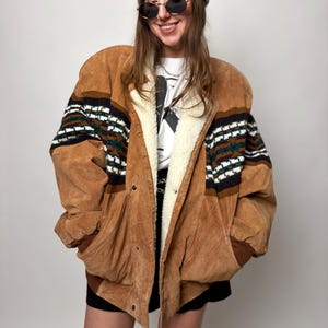 Op de afbeelding: Een bruine suède jas met een crèmekleurige shearling voering en een patroon op de mouwen. De jas heeft een knoopsluiting en zakken. De persoon draagt een grijze muts en zonnebril.