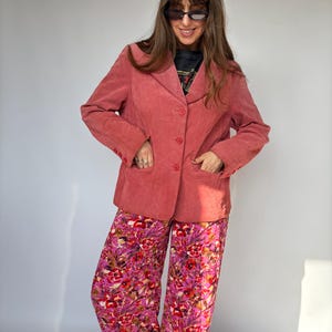 May include: A dusty rose corduroy blazer featuring a notched lapel, three front buttons, and long sleeves with buttoned cuffs, paired with wide-leg trousers showcasing a vibrant floral pattern in shades of pink, red, and purple.