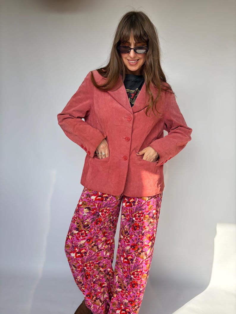 May include: A dusty rose corduroy blazer featuring a notched lapel, three front buttons, and long sleeves with buttoned cuffs, paired with wide-leg trousers showcasing a vibrant floral pattern in shades of pink, red, and purple.
