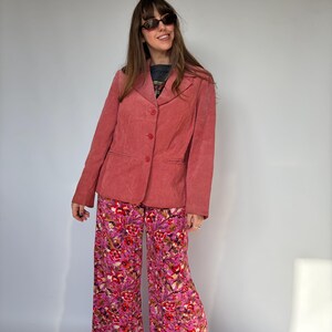 May include: A vintage-style outfit featuring a dusty rose blazer and wide-leg pants. The blazer has a three-button closure. The pants are covered in a vibrant floral pattern with shades of pink, red, and white. The outfit is completed with sunglasses.