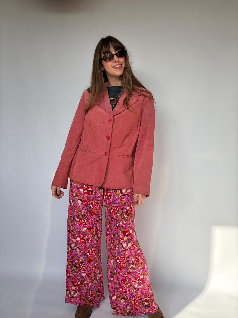 May include: A vintage-style outfit featuring a dusty rose blazer and wide-leg pants. The blazer has a three-button closure. The pants are covered in a vibrant floral pattern with shades of pink, red, and white. The outfit is completed with sunglasses.