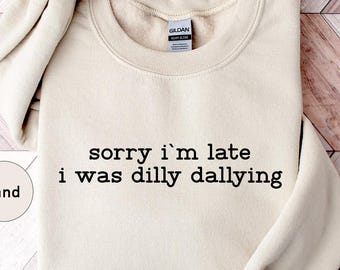 May include: A sand-colored sweatshirt with the text "sorry i'm late i was dilly dallying" in black. The sweatshirt is folded neatly on a light brown and white wooden surface. The word "Sand" is in a circle on the left.