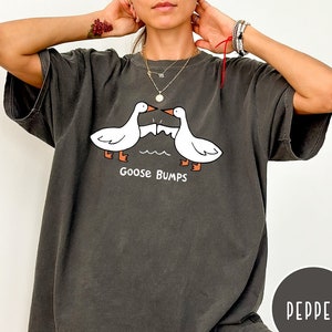 Może przedstawiać: Oversize'owy t-shirt w kolorze pieprzu z kresk&oacute;wkowym wzorem dw&oacute;ch białych gęsi z pomarańczowymi stopami i dziobami, zwr&oacute;conych do siebie. Pod gęsiami nadrukowano napis "Goose Bumps". Koszulka ma kr&oacute;tkie rękawy i luźny kr&oacute;j.
