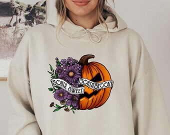 May include: A cream-colored hoodie with a graphic of a pumpkin with a spooky face and flowers. The pumpkin is split in half, with one side saying "Sorta Sweet" and the other side saying "Sorta Spooky".