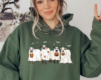 May include: A green hooded sweatshirt featuring a design of four sheep dressed as ghosts for Halloween. The sheep are holding pumpkins and are surrounded by bats and stars.