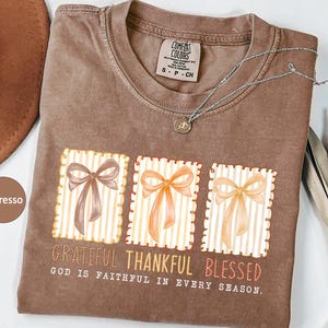 May include: Espresso-colored t-shirt with a graphic of three bows in frames, with the words "Grateful, Thankful, Blessed." The text "God is faithful in every season" is also printed on the shirt. A silver necklace and a brown hat are also visible.