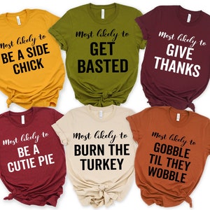 May include: A collection of six colorful t-shirts with humorous text. The shirts are in shades of gold, olive green, burgundy, beige, and rust. Each shirt has a different phrase in black script, such as "Most likely to be a side chick."