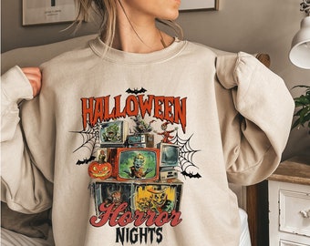 May include: A beige sweatshirt with a graphic print of a vintage television set with a Halloween theme. The graphic includes the text "Halloween Horror Nights" and features a variety of spooky characters and imagery.