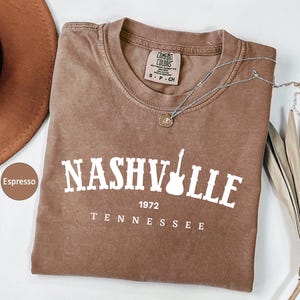 May include: Espresso-coloured t-shirt with the word "Nashville" and a guitar graphic. The text "1972 Tennessee" is below. A silver necklace with a pendant and a brown hat are also visible.