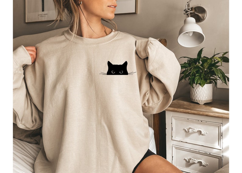 May include: A beige sweatshirt with a black cat peeking out from behind the pocket. The cat has white whiskers and large black eyes.