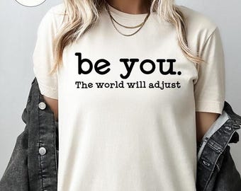 May include: Cream-colored t-shirt with the text "be you." above "The world will adjust" in black. The shirt is paired with a dark denim jacket. A small, circular "Natural" label is in the upper left corner.