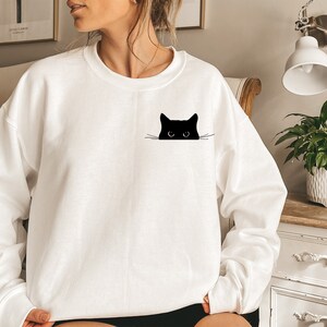 May include: White sweatshirt featuring a black cat peeking out from behind the fabric. The cat has large eyes and a simple design.