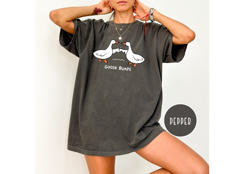 Może przedstawiać: Oversize'owy t-shirt w kolorze pieprzu z kresk&oacute;wkowym wzorem dw&oacute;ch białych gęsi z pomarańczowymi stopami i dziobami, zwr&oacute;conych do siebie. Pod gęsiami nadrukowano napis "Goose Bumps". Koszulka ma kr&oacute;tkie rękawy i luźny kr&oacute;j.
