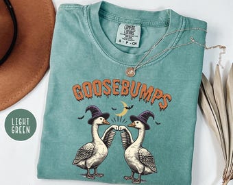 May include: Light green Comfort Colors t-shirt with a Halloween-themed graphic. The graphic features two geese wearing witch hats, with the word "GOOSEBUMPS" in orange. A crescent moon and bats are also included in the design.