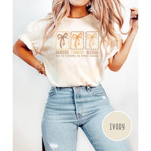 May include: Ivory-colored t-shirt with three bow designs in brown, orange, and tan. The text "Grateful Thankful Blessed" and "God is faithful in every season" is printed on the shirt. The model is wearing light blue jeans.