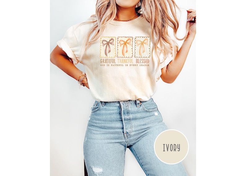 May include: Ivory-colored t-shirt with three bow designs in brown, orange, and tan. The text "Grateful Thankful Blessed" and "God is faithful in every season" is printed on the shirt. The model is wearing light blue jeans.