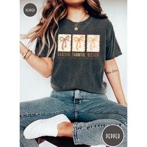 May include: Dark gray t-shirt with the words "Grateful Thankful Blessed" and three bow designs. The shirt is worn with blue jeans and white shoes. The overall theme is gratitude and blessings.