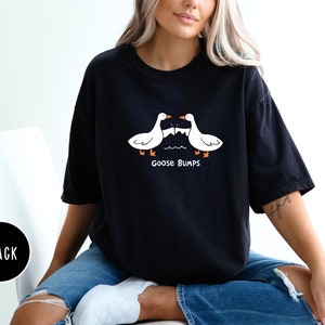 Może przedstawiać: Czarny t-shirt z fantazyjnym wzorem z dwoma białymi gęsiami zwr&oacute;conymi do siebie nad napisem "Goose Bumps". Gęsi mają pomarańczowe stopy i dzioby. Koszulka jest w swobodnym, wygodnym stylu.