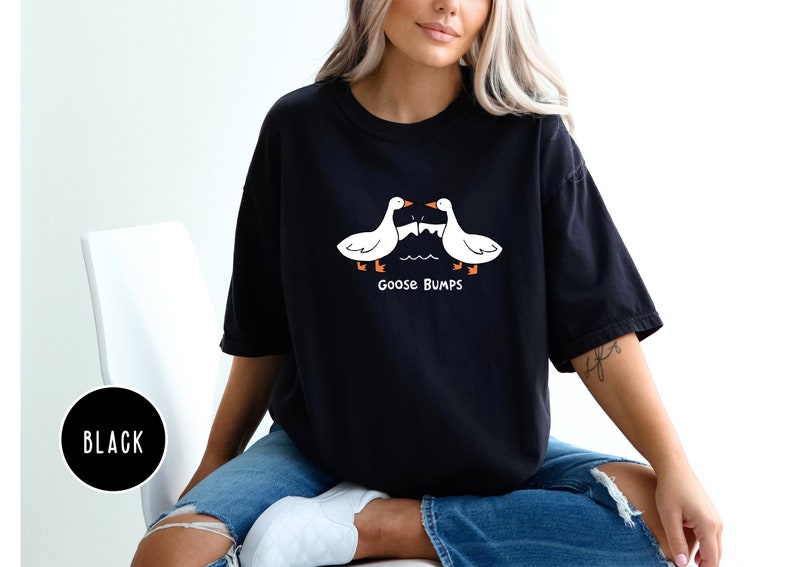 Może przedstawiać: Czarny t-shirt z fantazyjnym wzorem z dwoma białymi gęsiami zwr&oacute;conymi do siebie nad napisem "Goose Bumps". Gęsi mają pomarańczowe stopy i dzioby. Koszulka jest w swobodnym, wygodnym stylu.