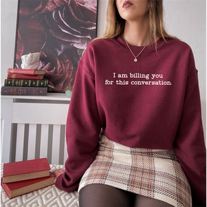May include: A maroon sweatshirt with the text "I am billing you for this conversation." in white lettering. The sweatshirt is worn by a person wearing a plaid skirt and black tights.