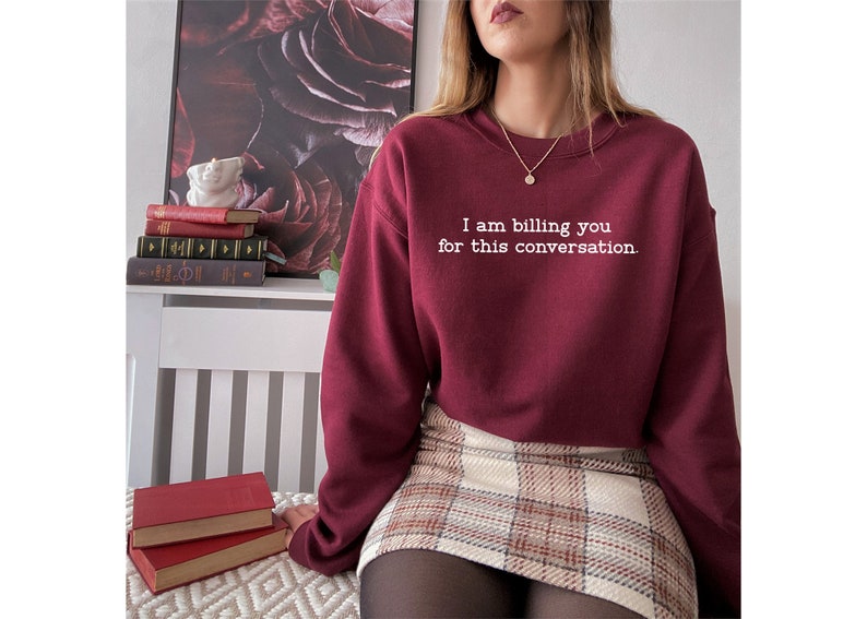 May include: A maroon sweatshirt with the text "I am billing you for this conversation." in white lettering. The sweatshirt is worn by a person wearing a plaid skirt and black tights.