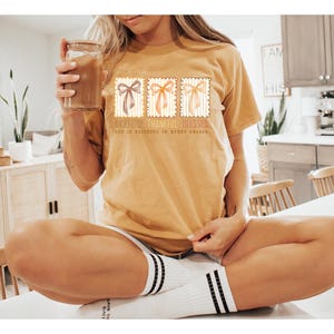 May include: Mustard yellow t-shirt with three postage stamp-style bow designs. The text on the shirt reads "Grateful, Thankful, Blessed. God is Faithful in Every Season." The shirt is worn with white socks with black stripes.