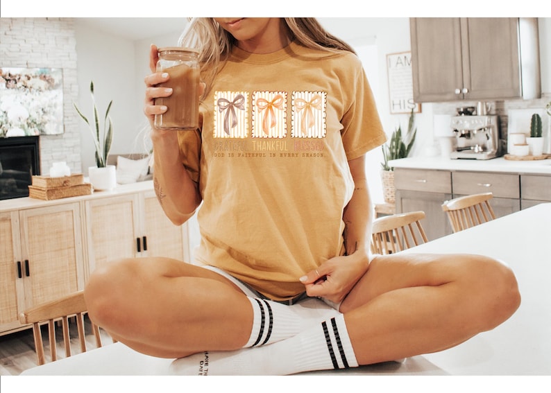 May include: Mustard yellow t-shirt with three postage stamp-style bow designs. The text on the shirt reads "Grateful, Thankful, Blessed. God is Faithful in Every Season." The shirt is worn with white socks with black stripes.