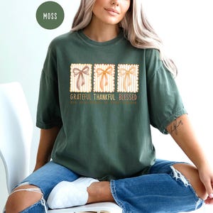 May include: Moss green t-shirt with the words "Grateful Thankful Blessed" and three bow illustrations. The shirt is paired with blue jeans. The text "God is faithful to those who trust" is also printed on the shirt.