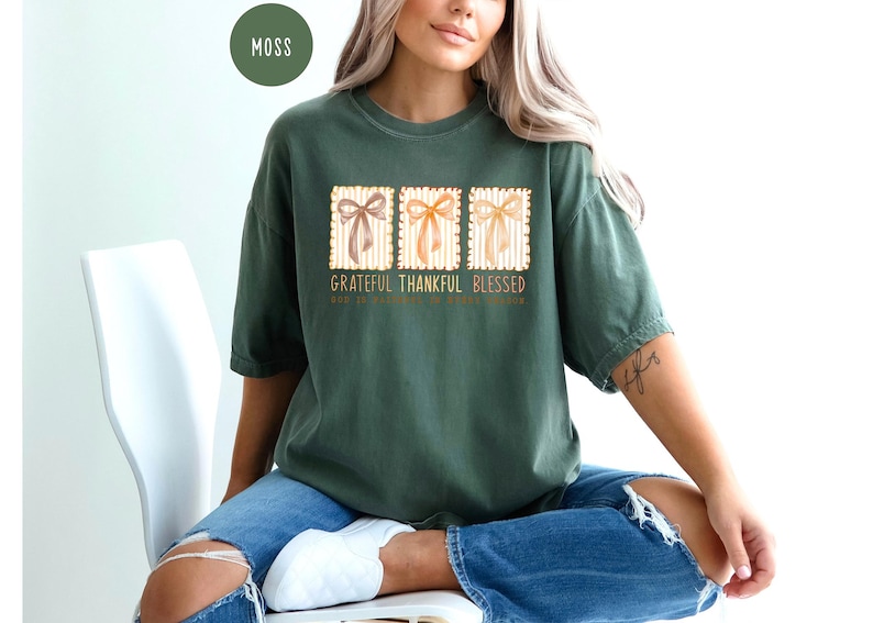 May include: Moss green t-shirt with the words "Grateful Thankful Blessed" and three bow illustrations. The shirt is paired with blue jeans. The text "God is faithful to those who trust" is also printed on the shirt.