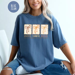 May include: A blue jean colored t-shirt with the words "Grateful Thankful Blessed" printed on the front. The design includes three rectangular images of bows. The shirt is a comfortable, casual style.