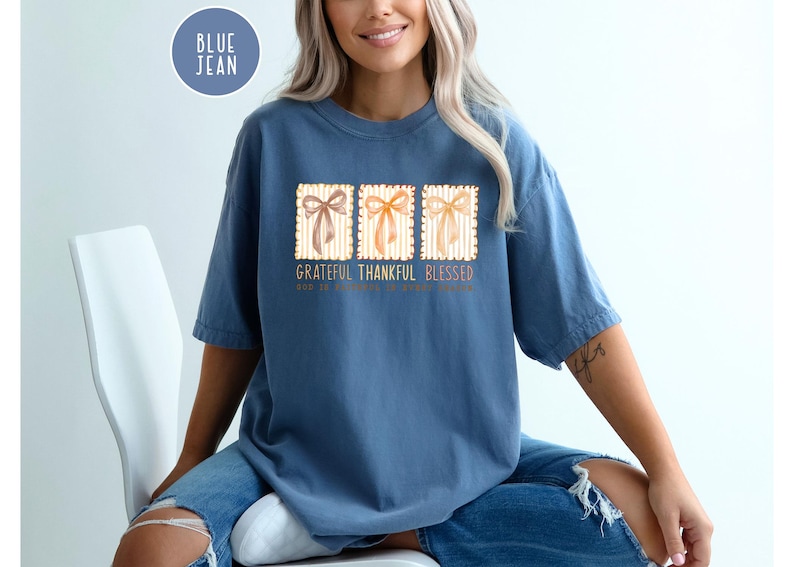 May include: A blue jean colored t-shirt with the words "Grateful Thankful Blessed" printed on the front. The design includes three rectangular images of bows. The shirt is a comfortable, casual style.
