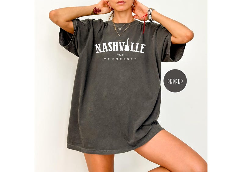 May include: Dark grey oversized t-shirt featuring the white text "NASHVILLE 1972 TENNESSEE" and a guitar graphic. The shirt has short sleeves and a loose fit, ideal for everyday wear.