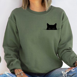 May include: A green sweatshirt with a black cat peeking out of a pocket. The cat has white whiskers and large eyes.