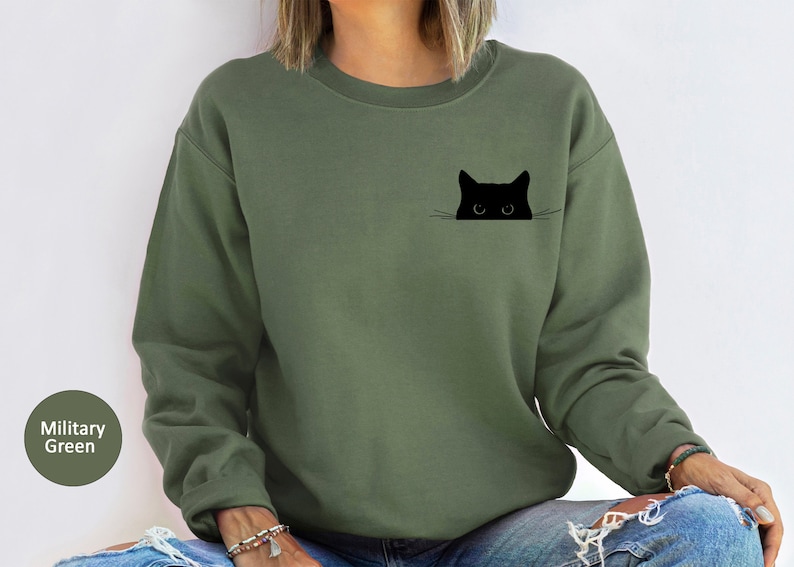 May include: A green sweatshirt with a black cat peeking out of a pocket. The cat has white whiskers and large eyes.