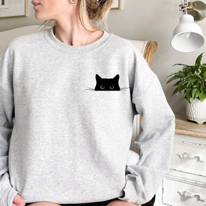 May include: A grey sweatshirt with a black cat peeking out from behind the pocket. The cat has white whiskers and large black eyes.