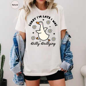 Może przedstawiać: Kremowy t-shirt z grafiką z kresk&oacute;wki przedstawiającą kaczkę. Kaczka biegnie z rozłożonymi skrzydłami, otoczona stokrotkami i napisem "Sorry I'm late I was Dilly Dallying". Koszulka jest połączona z dżinsową kurtką i czarnymi szortami.