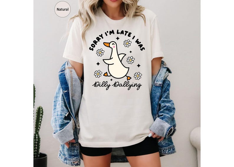 Może przedstawiać: Kremowy t-shirt z grafiką z kresk&oacute;wki przedstawiającą kaczkę. Kaczka biegnie z rozłożonymi skrzydłami, otoczona stokrotkami i napisem "Sorry I'm late I was Dilly Dallying". Koszulka jest połączona z dżinsową kurtką i czarnymi szortami.