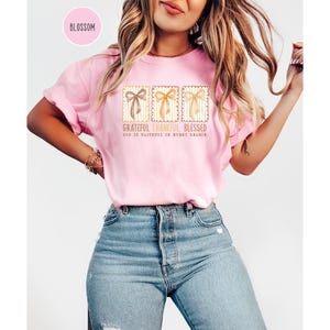 May include: A light pink t-shirt with a graphic of three decorative bows in a stamp-like design. The text on the shirt reads "Grateful, Thankful, Blessed" and "God is faithful in every season."