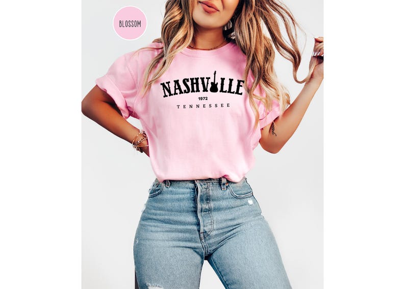 May include: A light pink t-shirt with the word "NASHVILLE" in black, a guitar silhouette, and the text "1972 TENNESSEE". The shirt is worn with blue jeans.
