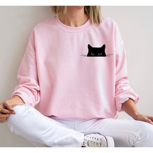 May include: A pink sweatshirt with a black cat peeking out from the pocket. The cat has large eyes and a small nose.